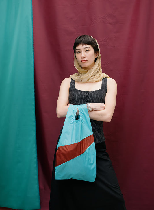 A model wearing a beige headscarf, a black ribbed tank top, and a black skirt holds a light blue tote bag with a diagonal brown stripe. The background is a split-color fabric backdrop with teal on the left and dark red on the right.
