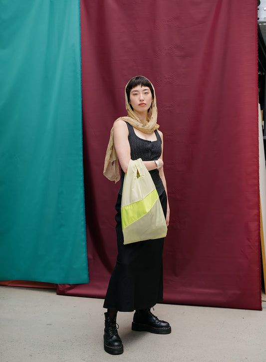 A model stands in front of a split teal and burgundy fabric backdrop, wearing a black ribbed tank top, black skirt, black boots, and a beige headscarf while holding a beige tote bag with a bright yellow-green accent panel.