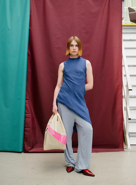 A model stands in front of a maroon and teal fabric backdrop, wearing a sleeveless blue mock-neck top, light gray wide-leg pants, and red shoes, while holding a beige tote bag with a pink stripe.
