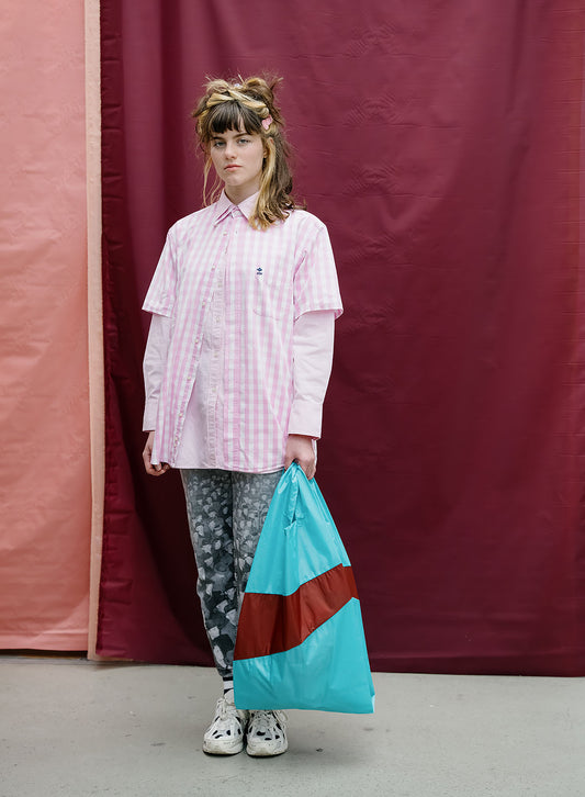 A model stands in front of a maroon and pink fabric backdrop, wearing a pink and white gingham button-up shirt, gray patterned leggings, and white sneakers, while holding a large turquoise and brown shopping bag.