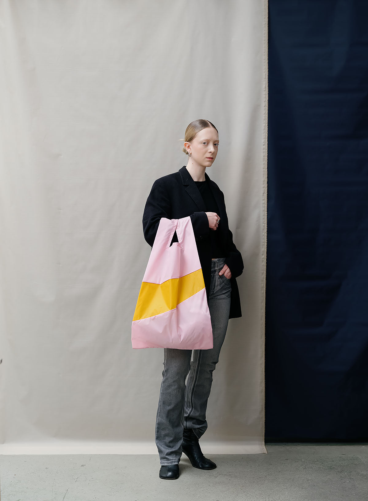 A model stands against a split beige and navy backdrop, holding a large pink tote bag with a yellow geometric stripe.