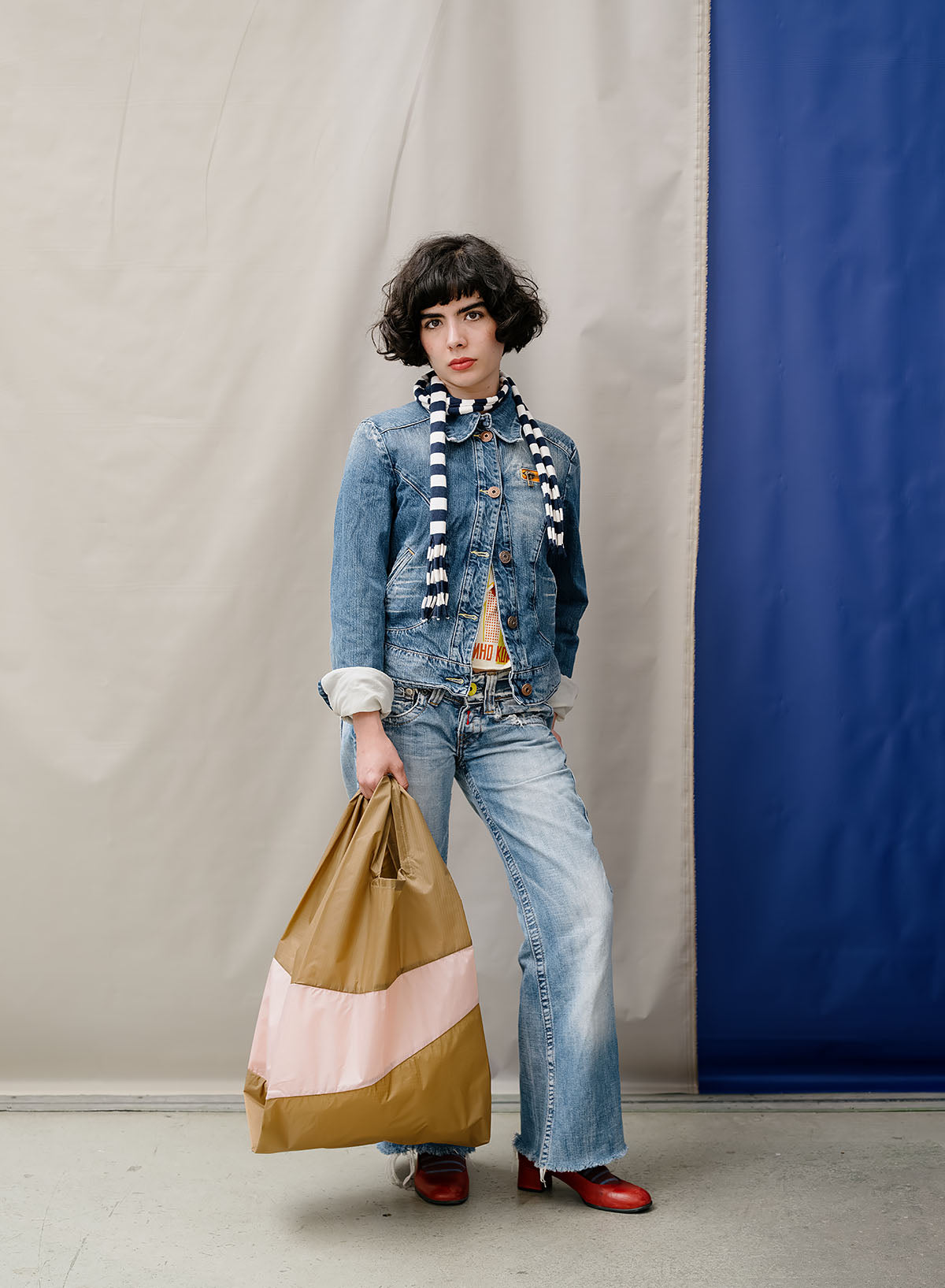 A model stands in a studio wearing a distressed blue denim jacket, matching wide-leg jeans, a black and white striped knit scarf, and red shoes, while holding a brown, pink, and beige striped paper shopping bag.