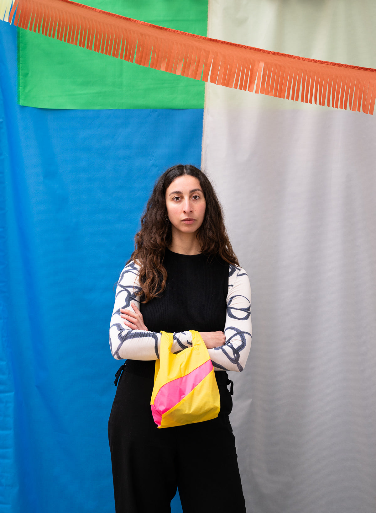 Certainly! Here’s your revised description, specifying the base and stripe colours: A woman with long, wavy hair stands with crossed arms, holding The New Shopping Bag in TV Yellow (base colour) & Fluo Pink (stripe colour), Small size, in front of blue, green, white, and orange backdrops—perfect for carrying daily essentials.