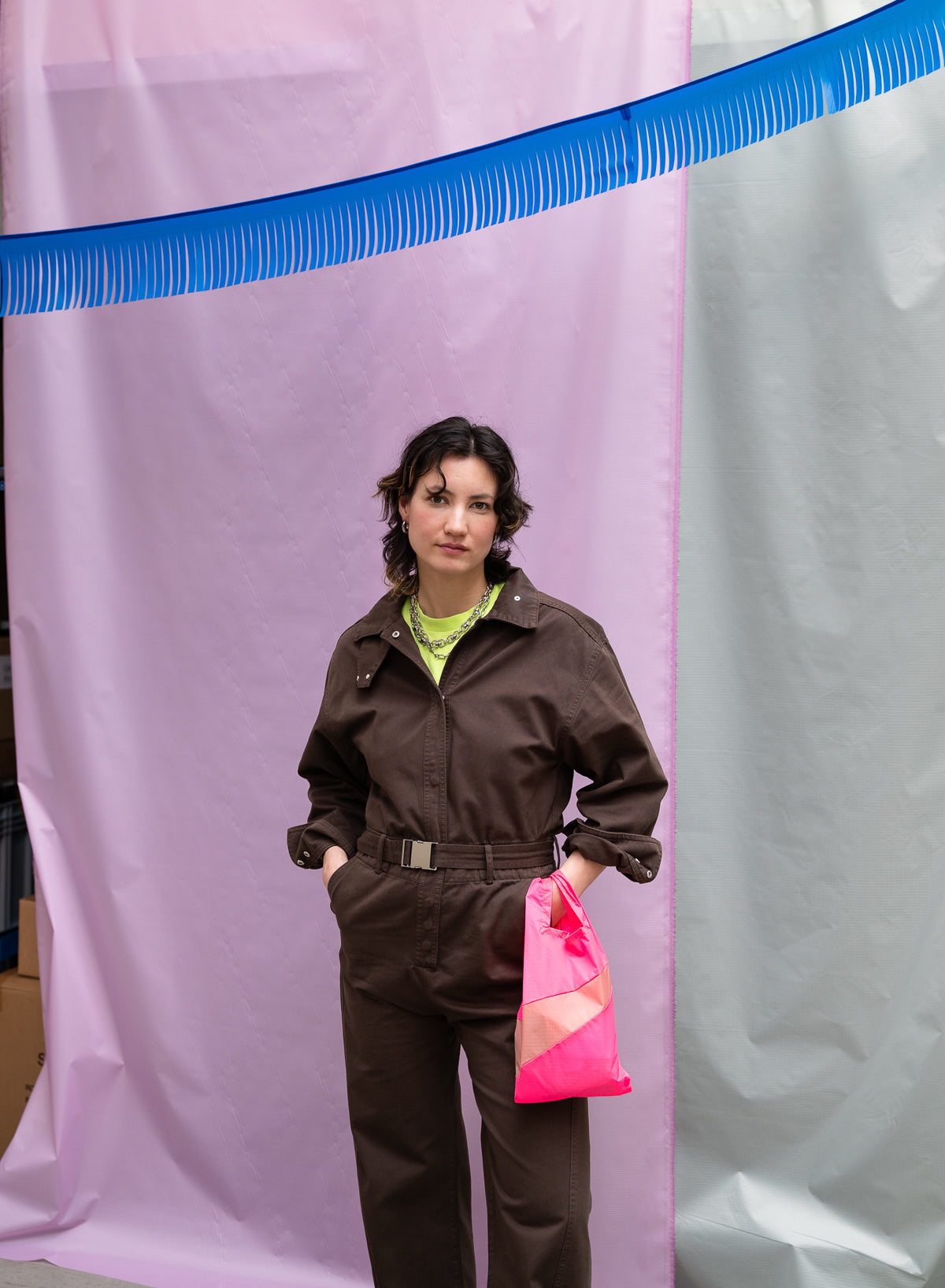 Certainly! Here’s your revised description, with clear mention of the base and stripe colours: A person with The New Shopping Bag Fluo Pink & Try Small (base colour: fluo pink, stripe colour: yellow) stands in front of pastel pink and gray curtains, a blue decorative element overhead, while wearing a brown jumpsuit.