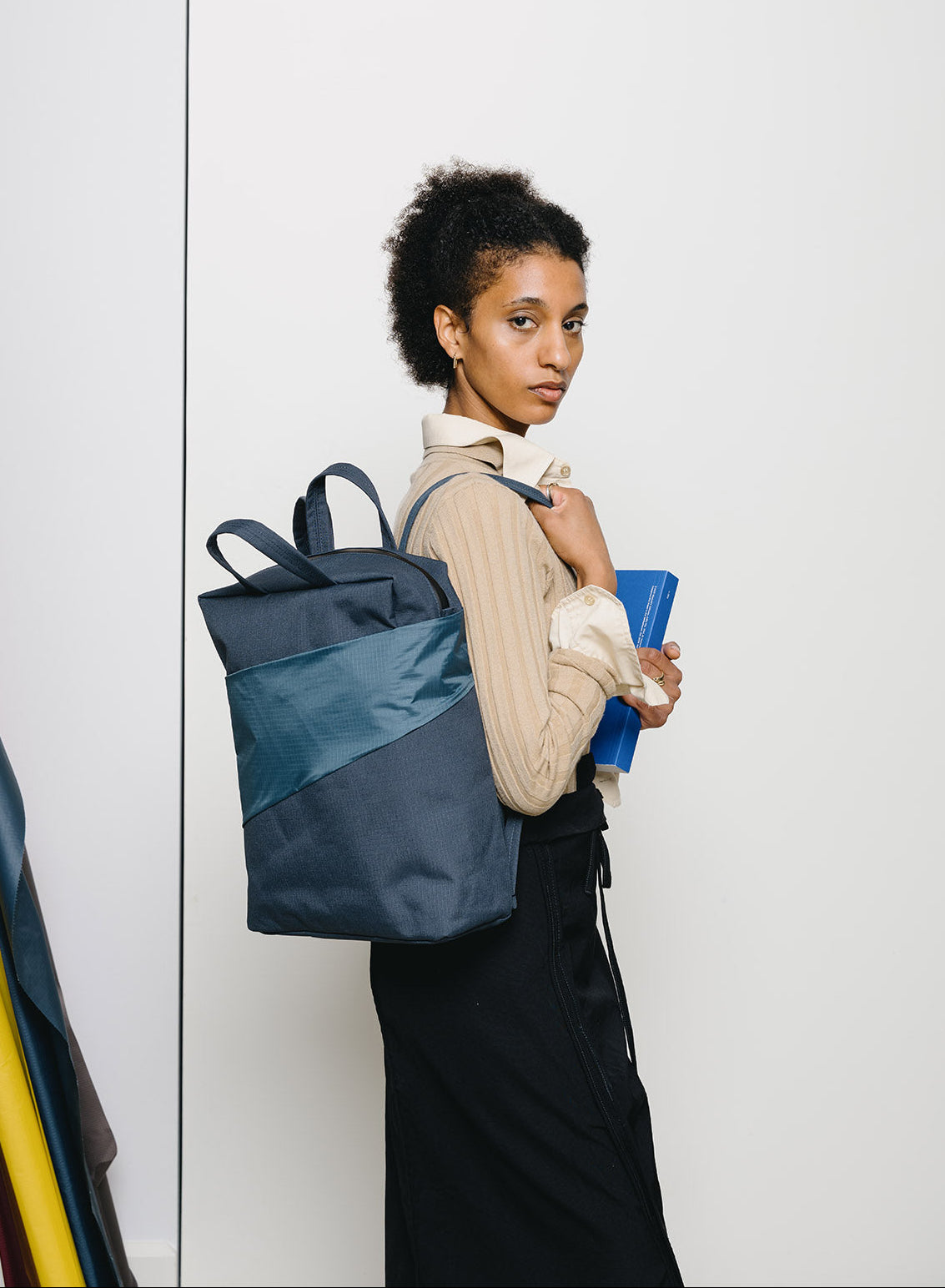 A woman with short curly dark hair stands against a white wall wearing a navy and slate blue color-blocked backpack, a beige ribbed long-sleeve top, and a black skirt, while holding a blue notebook.