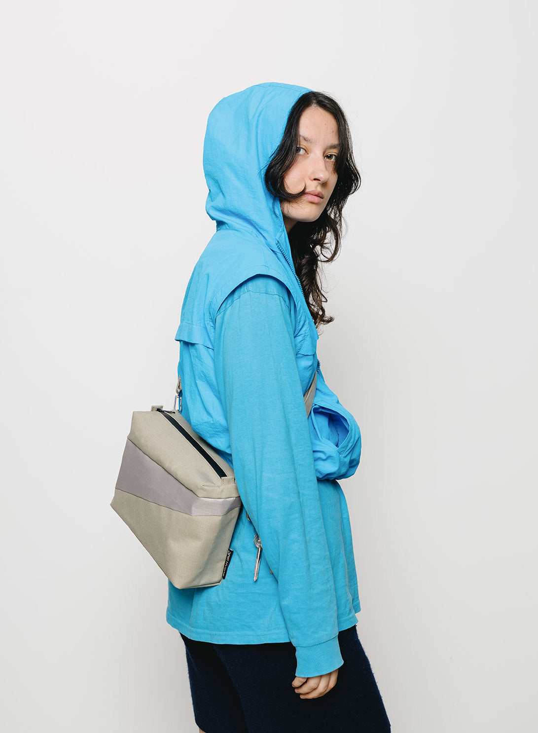 A model wearing a bright blue hooded windbreaker jacket and dark navy shorts, carrying a beige and gray geometric shoulder bag, set against a plain white studio background.