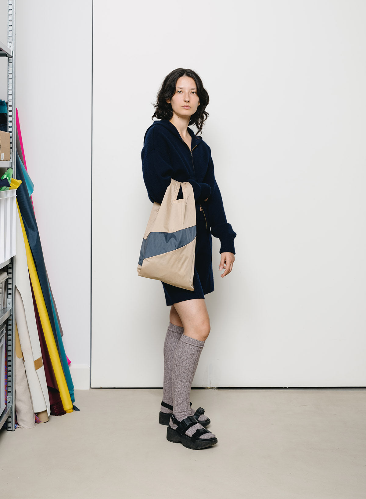 A woman stands in a minimalist studio setting wearing a navy blue long-sleeve shirt dress, gray knee-high socks, and black platform sandals, while carrying a beige tote bag with blue and gray color-blocked panels.