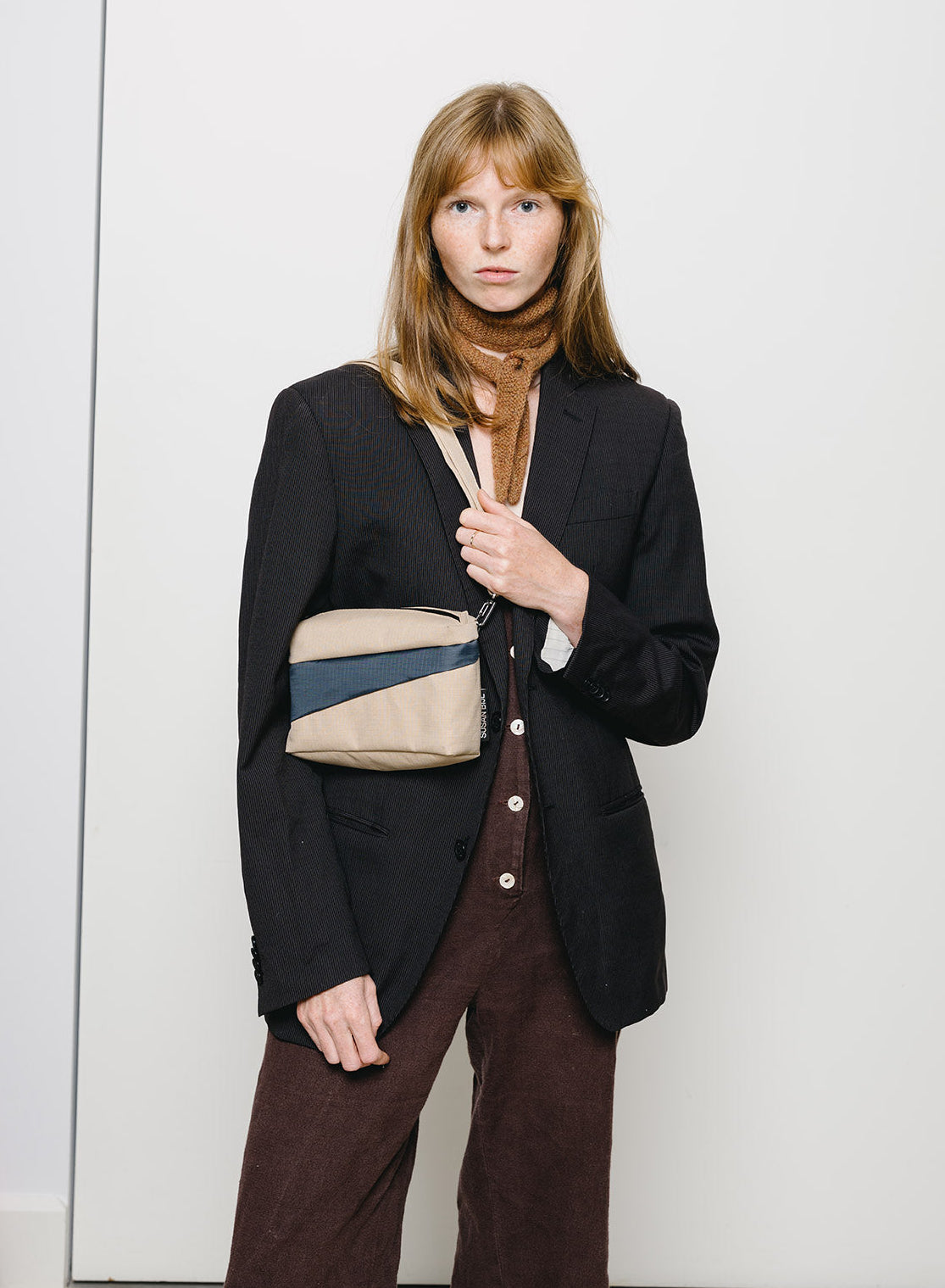 A model wearing a black blazer, brown corduroy trousers, and a brown scarf, carrying a beige and navy blue diagonal-striped crossbody bag against a plain white wall.
