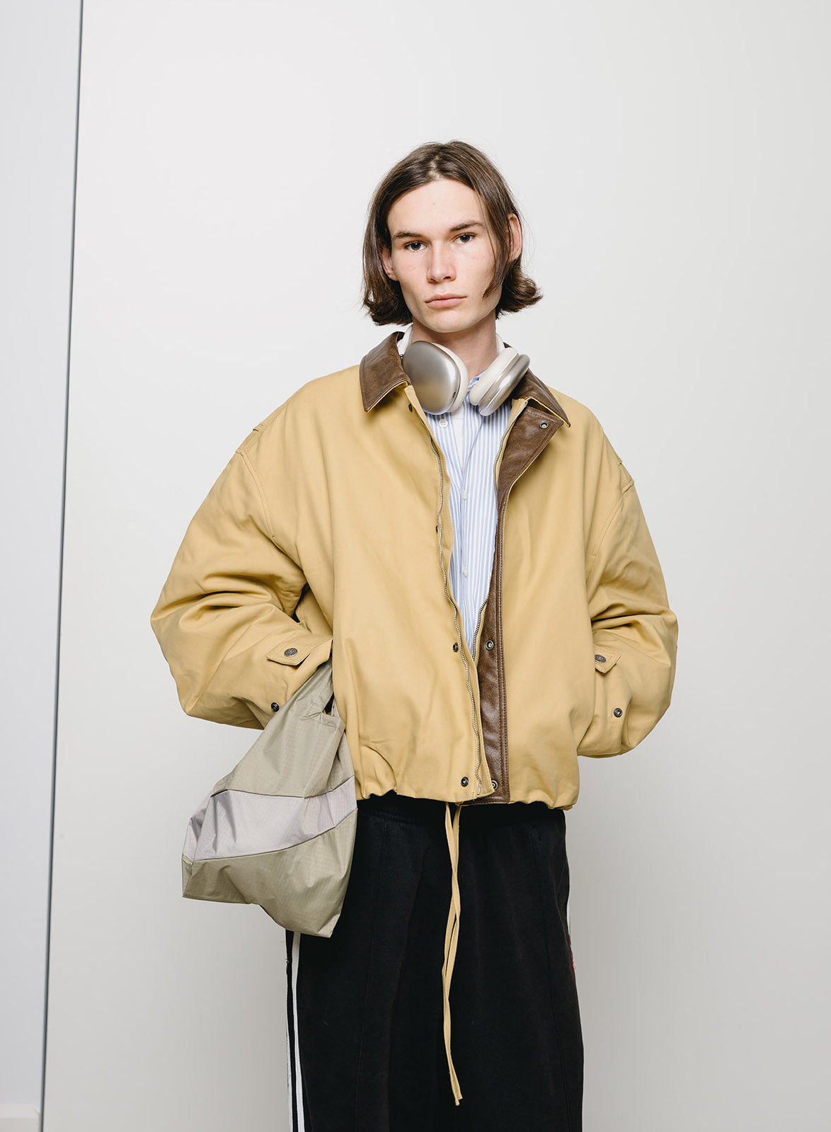 A model stands against a light gray studio wall wearing a cropped tan jacket with brown collar and sleeve trim, a white striped shirt, black pants, and carrying a beige and gray bag.