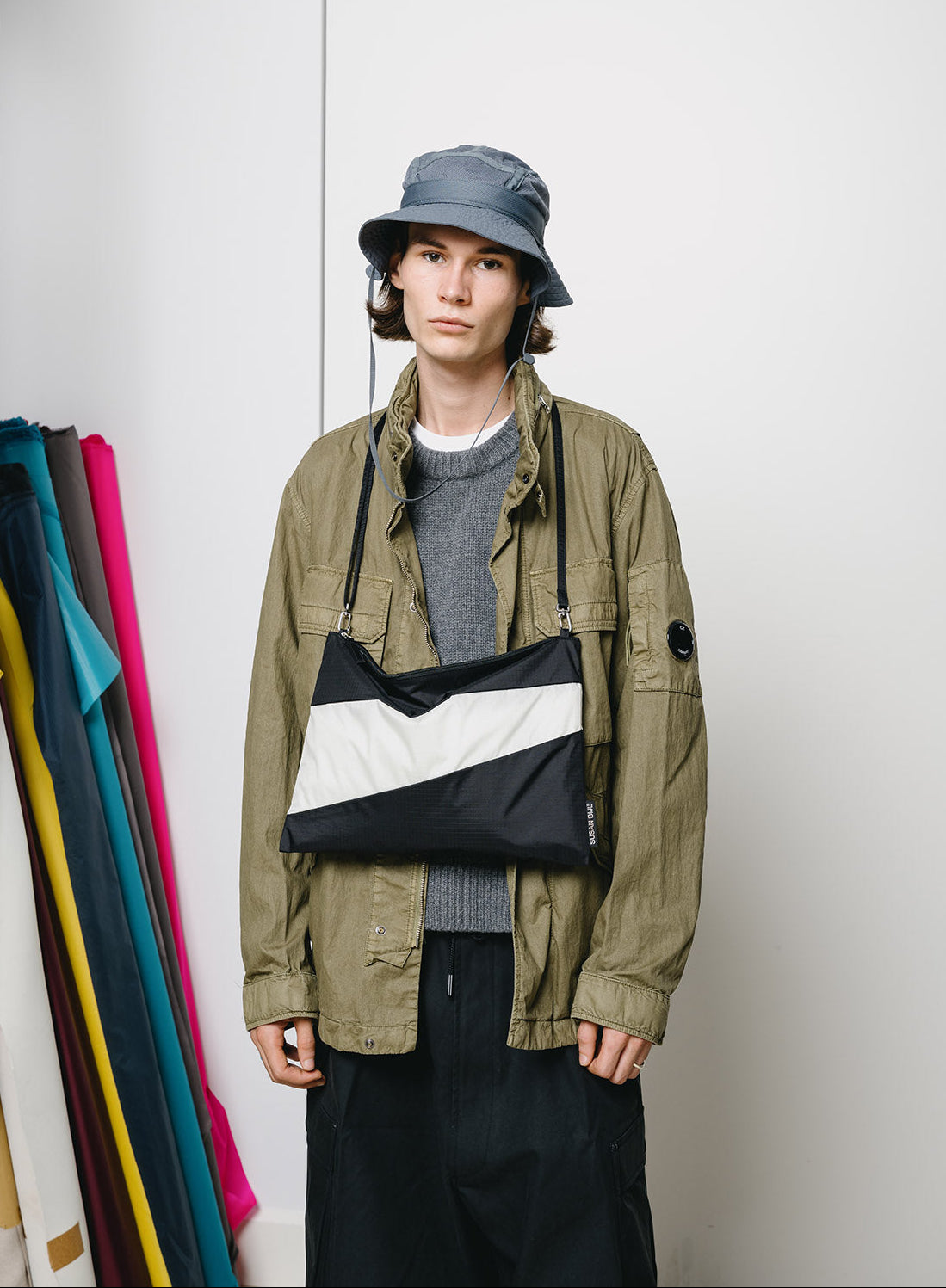 A model wearing an olive green utility jacket, gray knit sweater, black pants, and a blue-gray bucket hat, carrying a black and white diagonal-striped shoulder bag across their chest in a studio setting.
