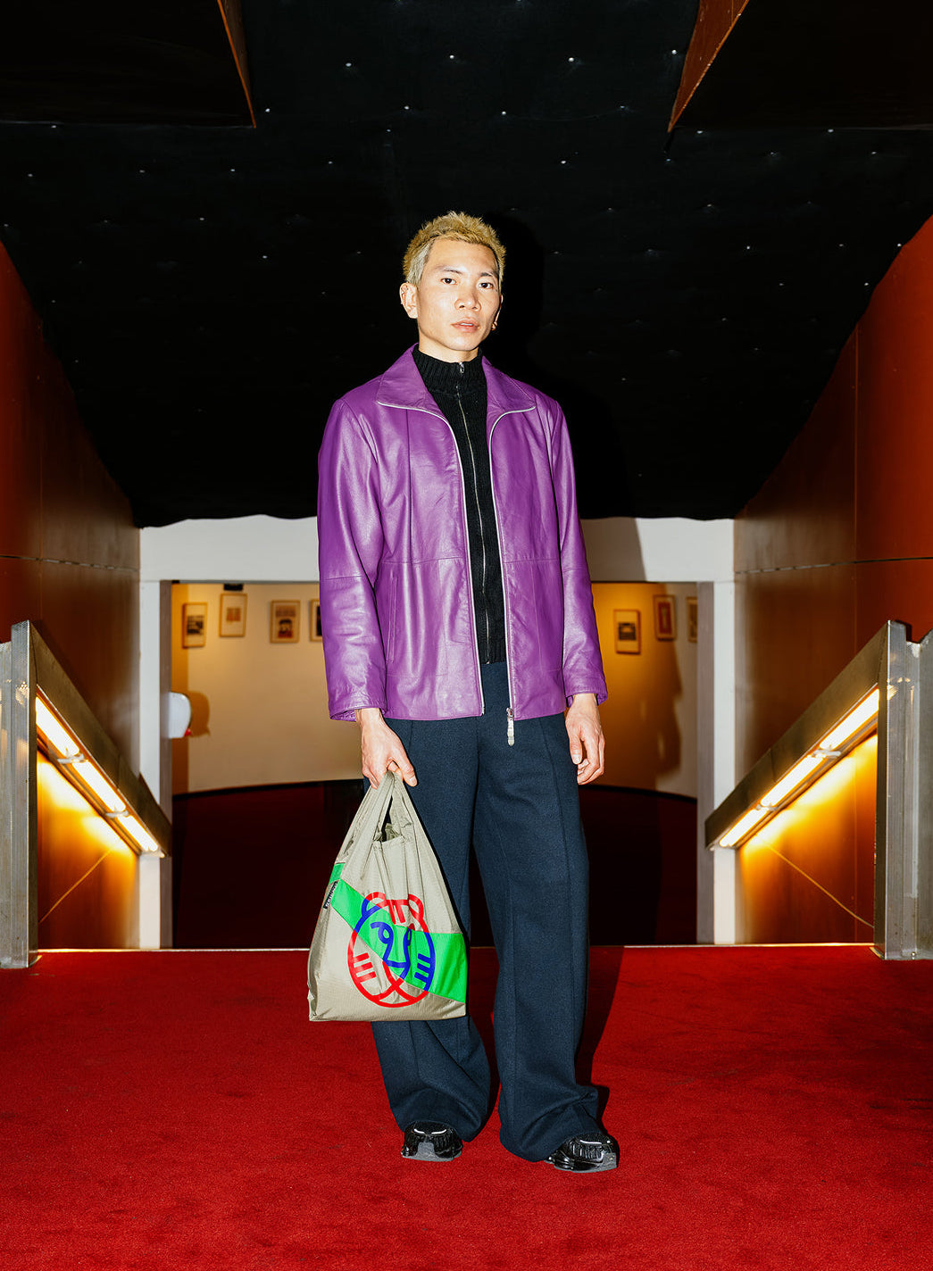Person wearing a purple jacket and dark pants standing on a red carpet with a decorative ceiling.