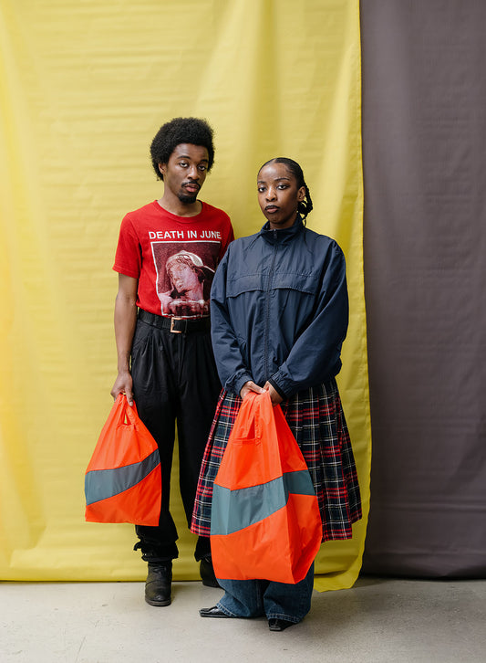 Two models standing in front of a split yellow and dark gray backdrop, holding bright orange and gray reflective bags. The model on the left wears a red graphic t-shirt and black pants, while the model on the right wears a navy blue high-collar jacket over a red and black plaid skirt.