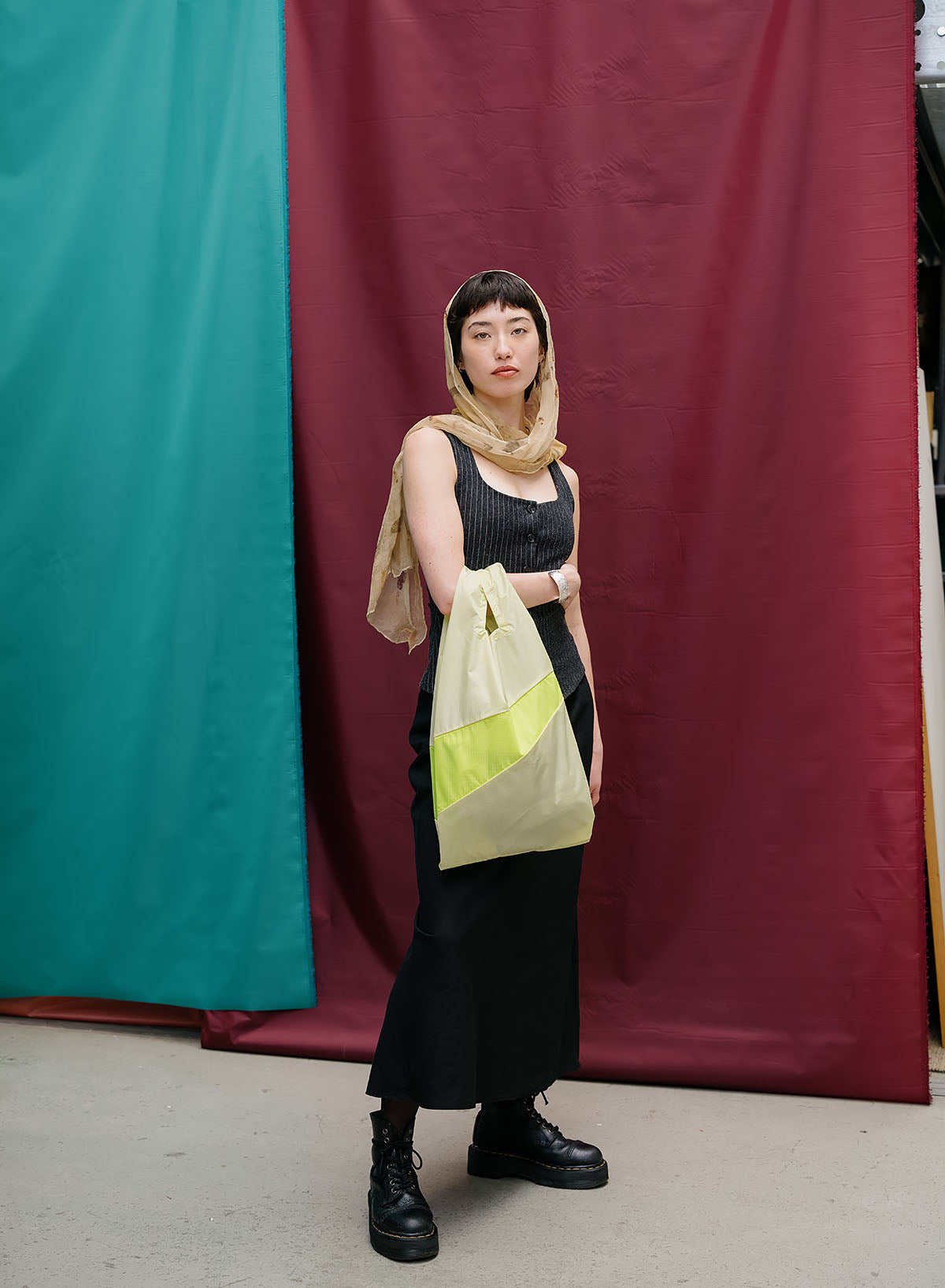 A model stands in front of a split teal and burgundy fabric backdrop, wearing a black ribbed tank top, black skirt, black boots, and a beige headscarf while holding a beige tote bag with a bright yellow-green accent panel.