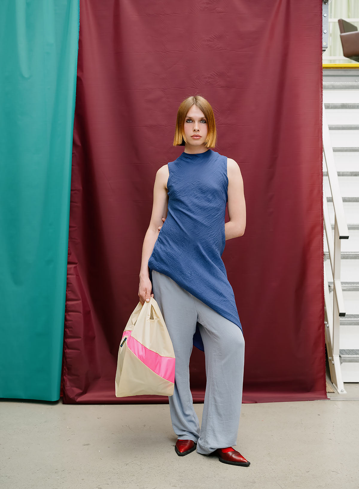 A model stands in front of a maroon and teal fabric backdrop, wearing a sleeveless blue mock-neck top, light gray wide-leg pants, and red shoes, while holding a beige tote bag with a pink stripe.
