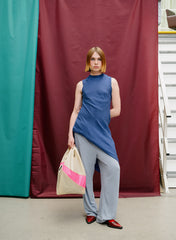 A model stands in front of a maroon and teal fabric backdrop, wearing a sleeveless blue mock-neck top, light gray wide-leg pants, and red shoes, while holding a beige tote bag with a pink stripe.