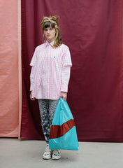 A model stands in front of a maroon and pink fabric backdrop, wearing a pink and white gingham button-up shirt, gray patterned leggings, and white sneakers, while holding a large turquoise and brown shopping bag.