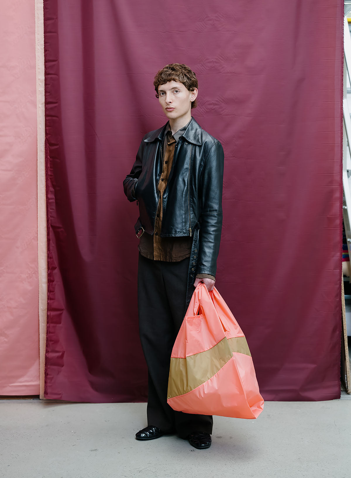A model stands in front of a burgundy and pink fabric backdrop, wearing a black leather jacket, brown shirt, and black trousers, while holding a large orange and beige striped plastic bag.