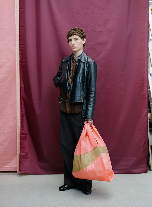 A model stands in front of a burgundy and pink fabric backdrop, wearing a black leather jacket, brown shirt, and black trousers, while holding a large orange and beige striped plastic bag.