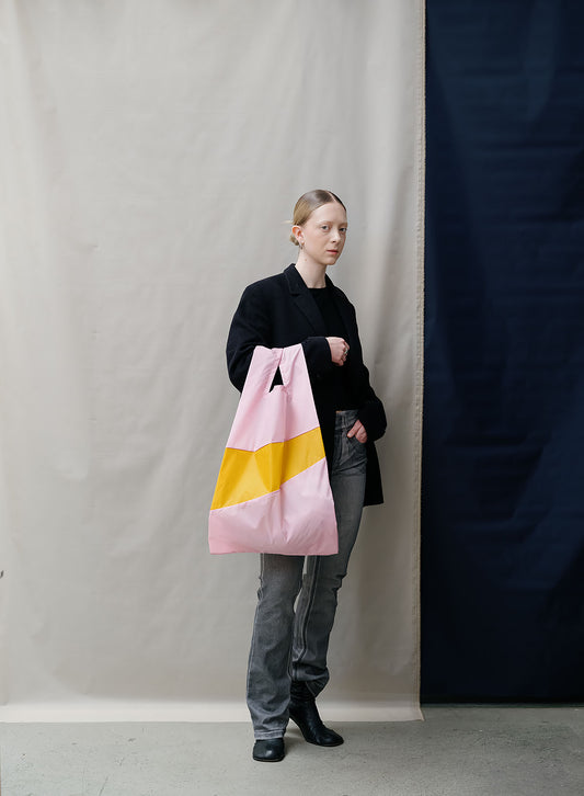 A model stands against a split beige and navy backdrop, holding a large pink tote bag with a yellow geometric stripe.