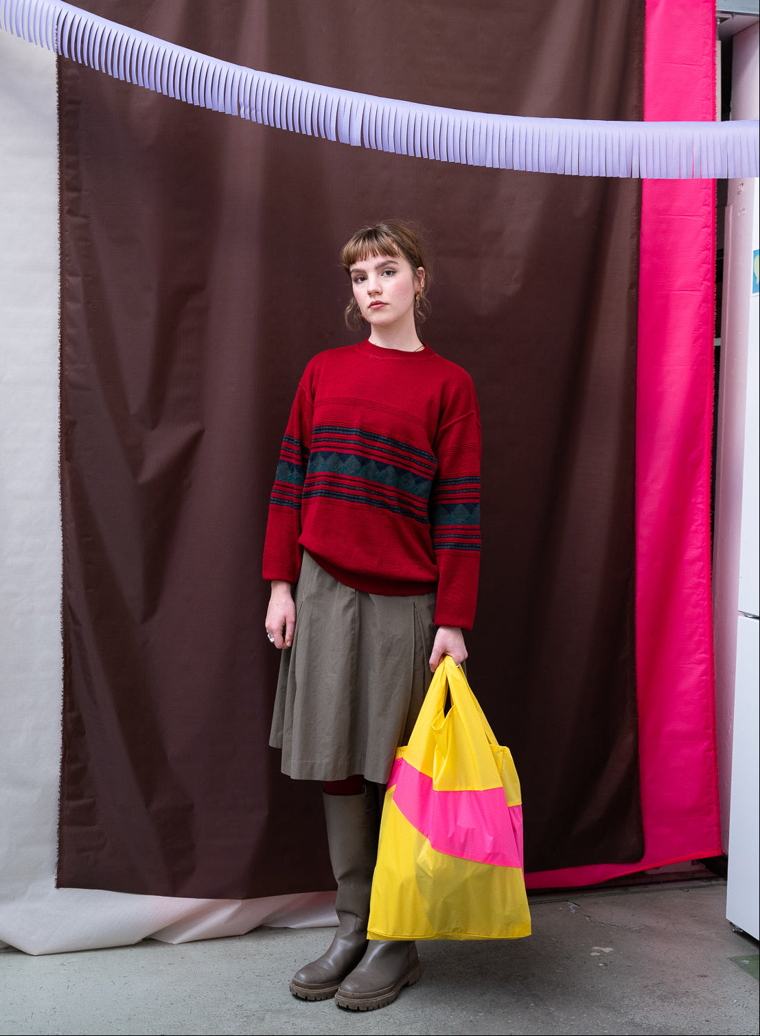 Person wearing a red sweater with a striped pattern, holding a yellow and pink bag, standing in front of a colorful curtain.