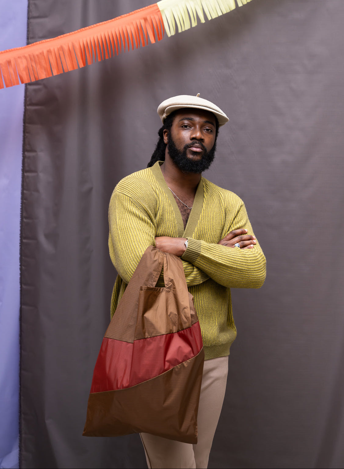 Man in a green sweater holding a brown bag against a colorful fringe backdrop