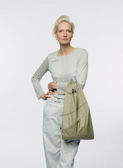 A woman with short blonde hair stands against a light gray studio background, wearing a light gray long-sleeve top and pale blue jeans while carrying a large olive green tote bag.