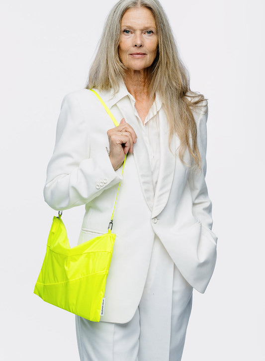 A woman with long blonde hair wearing a white blazer and trousers, holding a bright neon yellow-green shoulder bag against a plain light gray studio background.