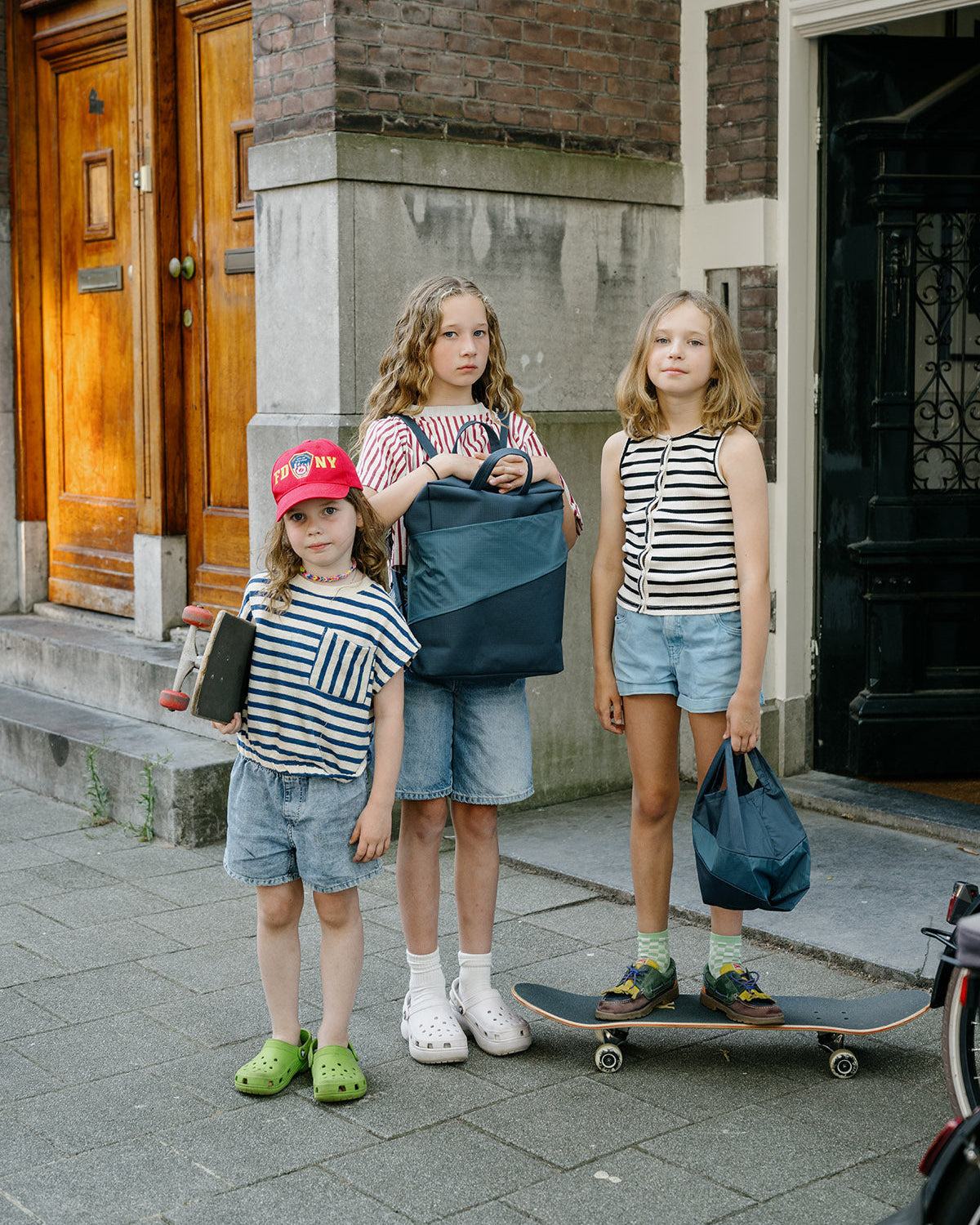 Three children standing on a city sidewalk in front of a brick building, each holding a navy blue tote bag. The children are wearing casual summer outfits including striped tank tops, denim shorts, and colorful sneakers.