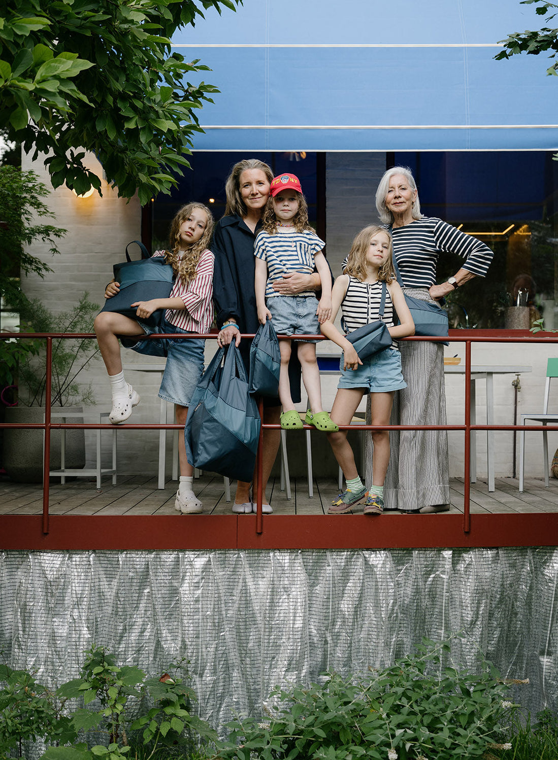 A family of five poses together on a wooden deck in front of a modern house. The group is dressed in casual summer clothing, and several of them are holding dark blue designer handbags.