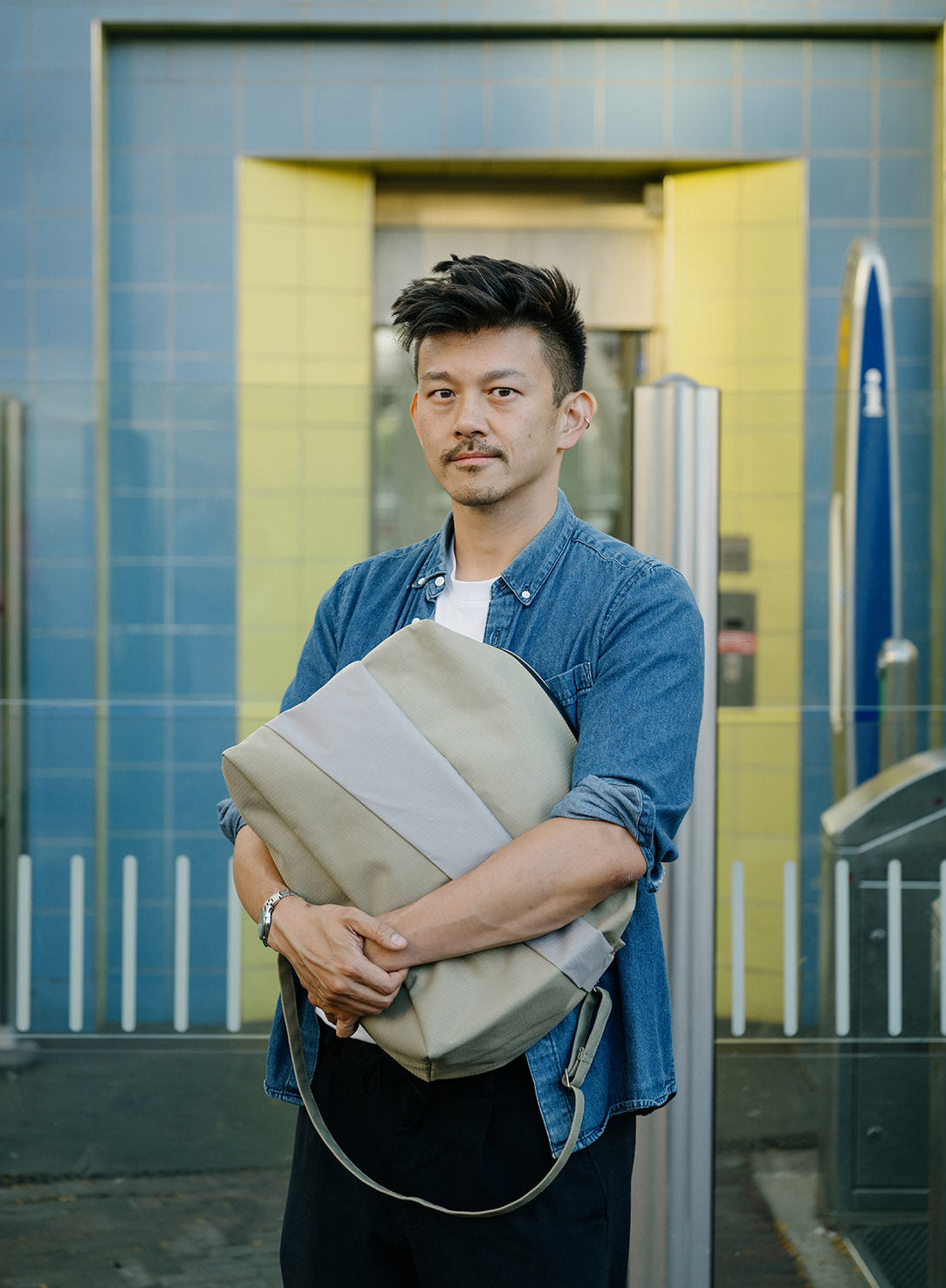 A man wearing a blue denim shirt and black pants stands in front of a blue tiled building, holding a large beige and gray shoulder bag.