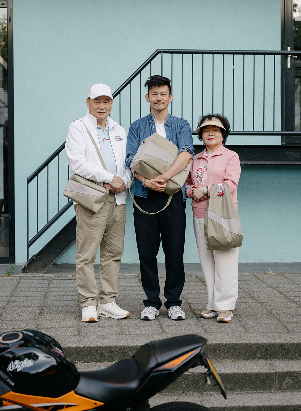 Three people standing on outdoor steps in front of a light blue building, each holding a beige tote bag. A black and orange motorcycle is visible in the foreground.